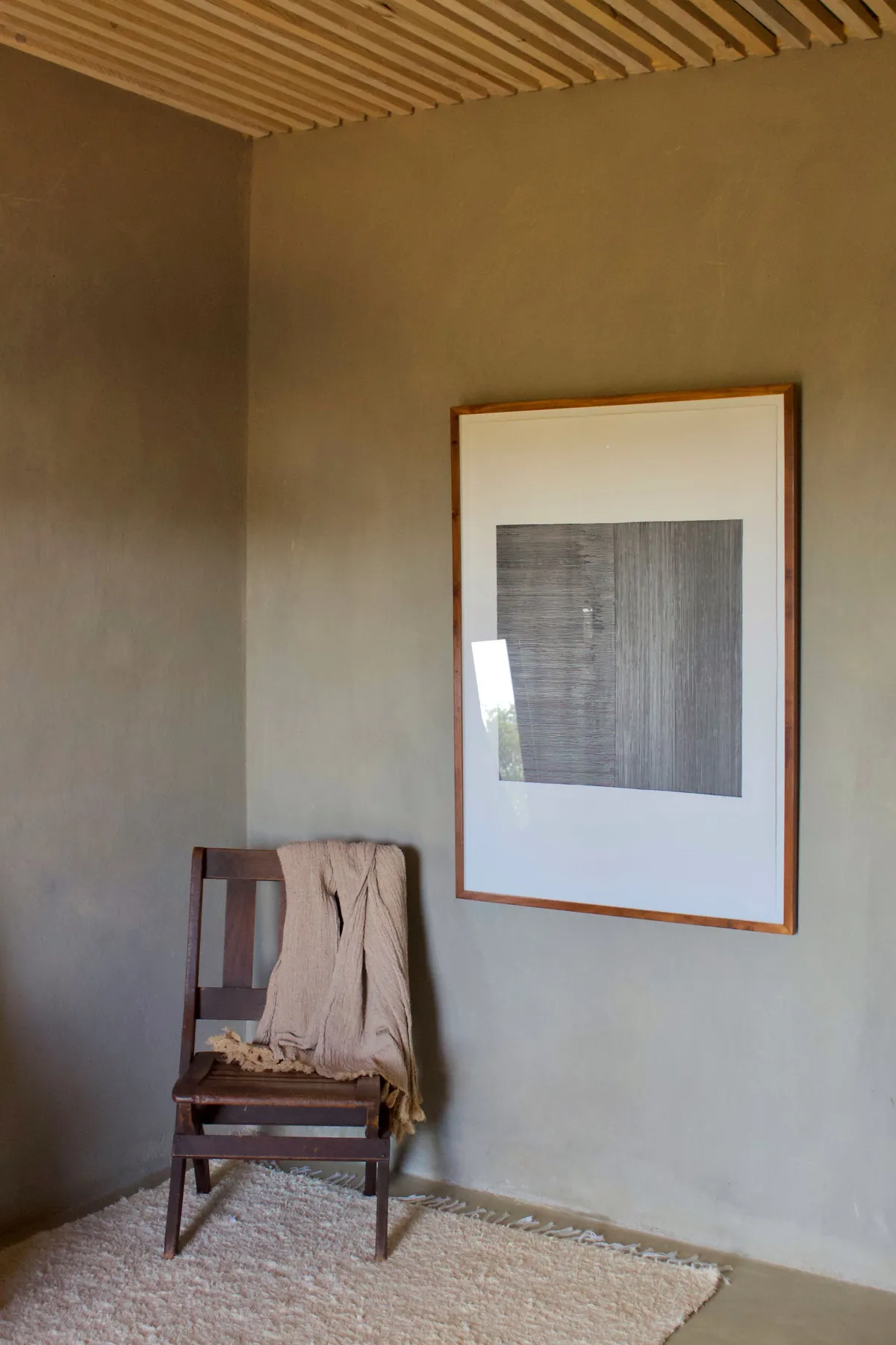 Plaster-walled corner with timber slat ceiling, wooden chair and framed artwork, daylight